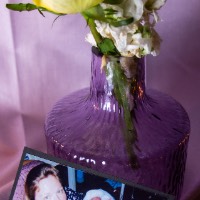 Photo of a photograph of a mother holding a baby. The photo sits on a purple cloth against a purple vase holding a yellow flower.